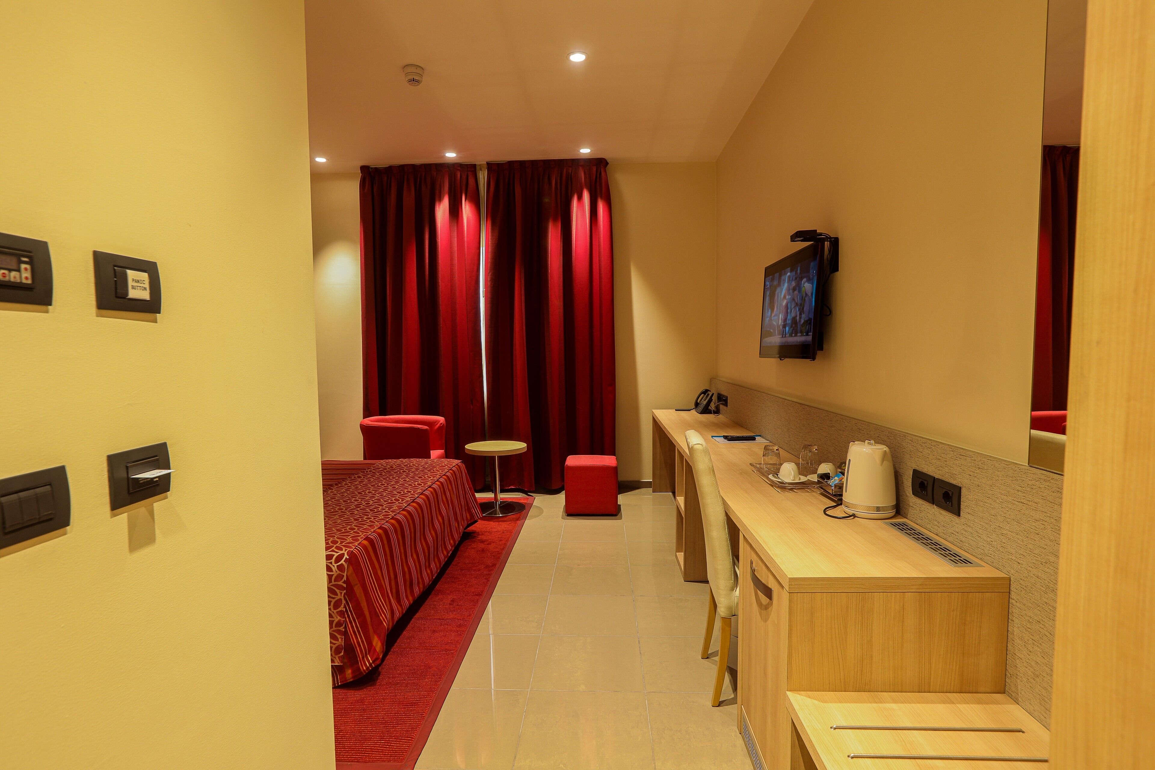 Habitación de hotel con cama, escritorio, silla, mesita pequeña, cortina roja y televisor montado en la pared.