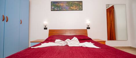 A hotel room with a red bedspread, a wooden headboard, a painting on the wall, and a blue wardrobe.