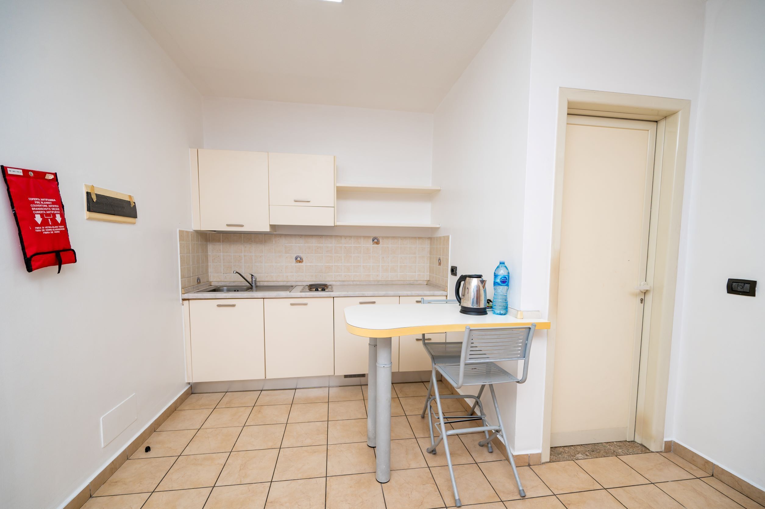 Una cocina compacta con muebles blancos, un lavamanos y una mesa con un hervidor y una botella de agua.