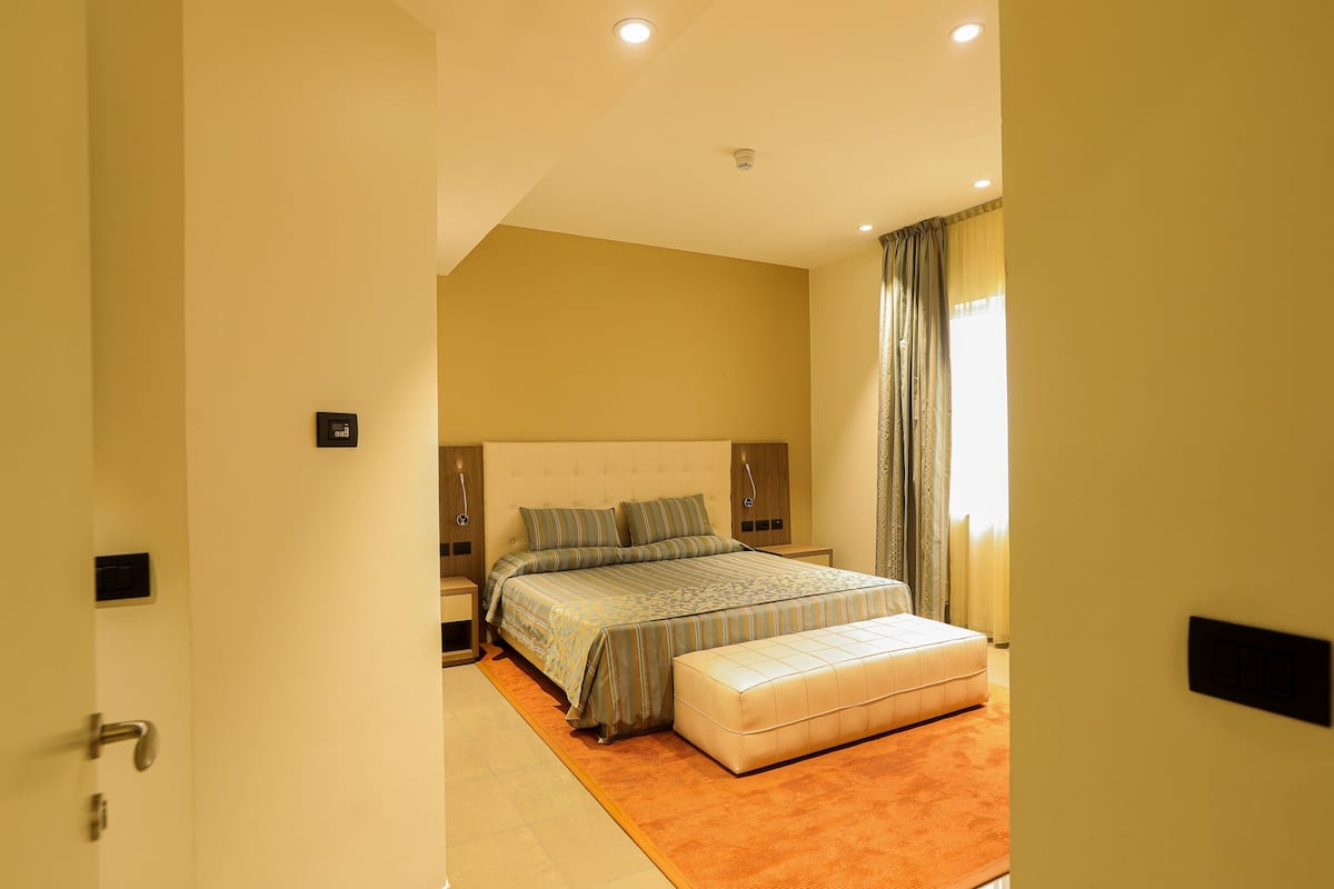 Une chambre à coucher avec un grand lit, une table de chevet, une commode et une fenêtre avec des rideaux.