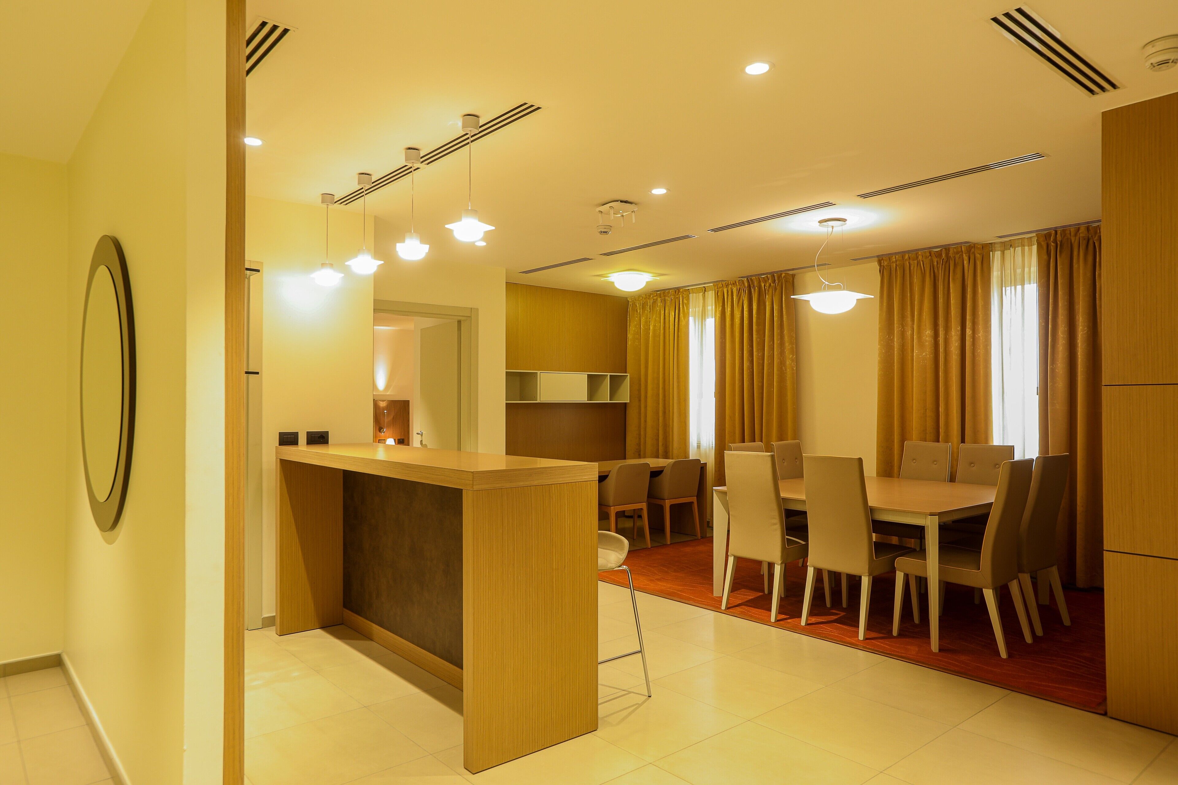 A modern hotel room with a wooden reception desk, a dining area with a table and chairs, and a kitchen in the background.