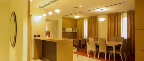 A modern hotel room with a wooden reception desk, a dining area with a table and chairs, and a kitchen in the background.