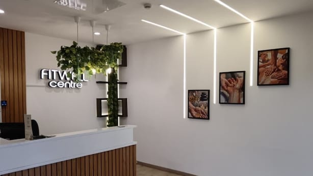 A reception area with a white reception desk, a sign reading 'FITWELL centre,' and framed pictures on the wall.