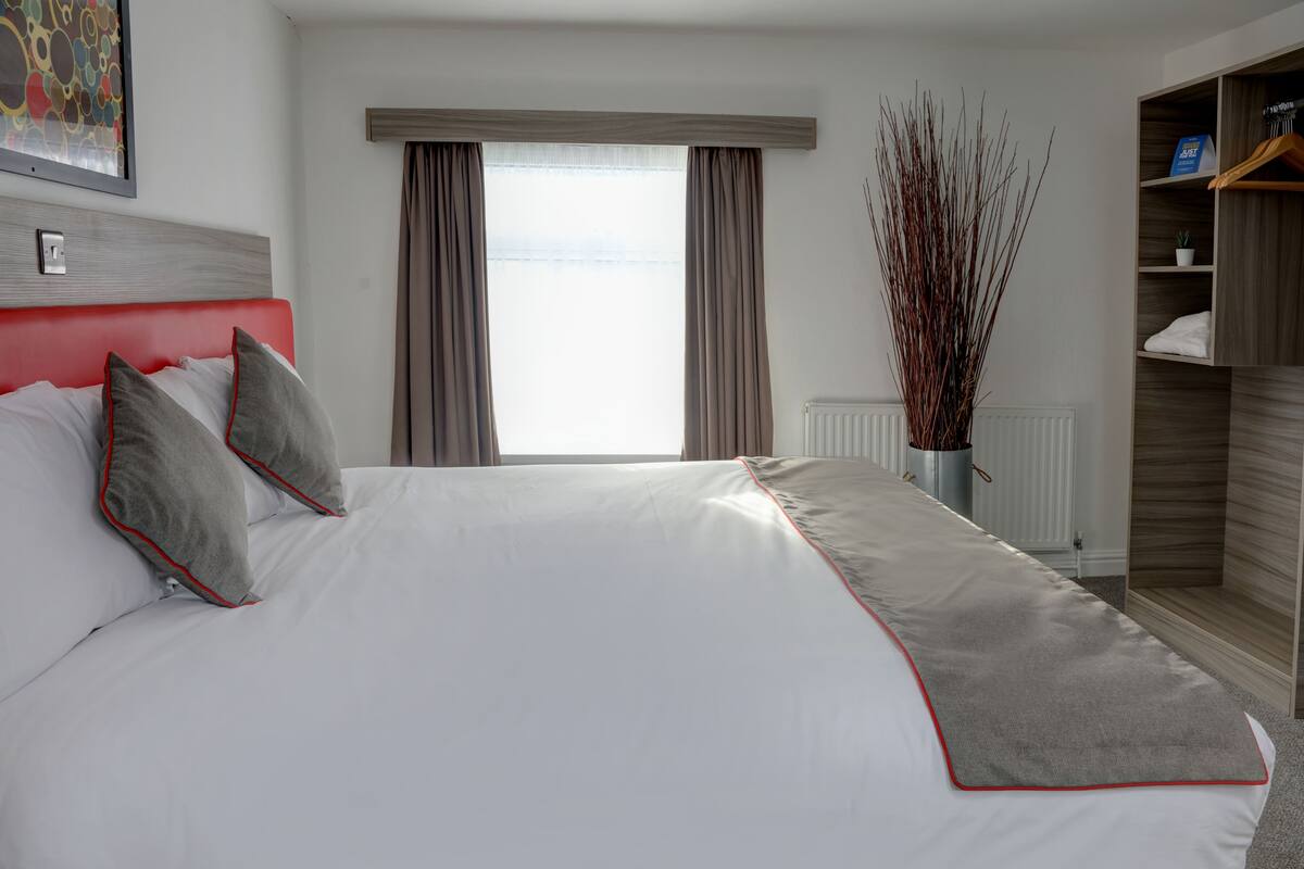 Una cama bien hecha con ropa de cama blanca y gris, un cabecero rojo y una ventana con cortinas.