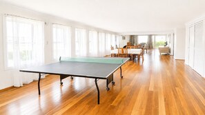 A spacious room with a ping pong table, wooden flooring, and large windows.