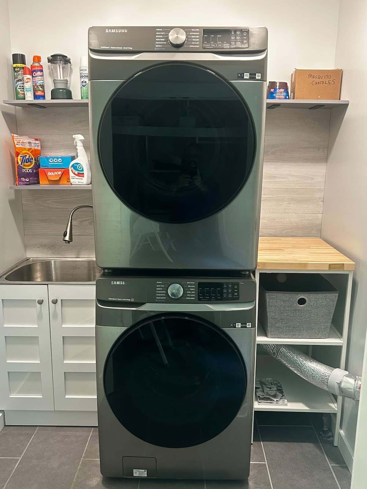 A stacked washer and dryer unit in a modern kitchen with a stainless steel sink, organized shelves with cleaning supplies, and a wooden countertop.