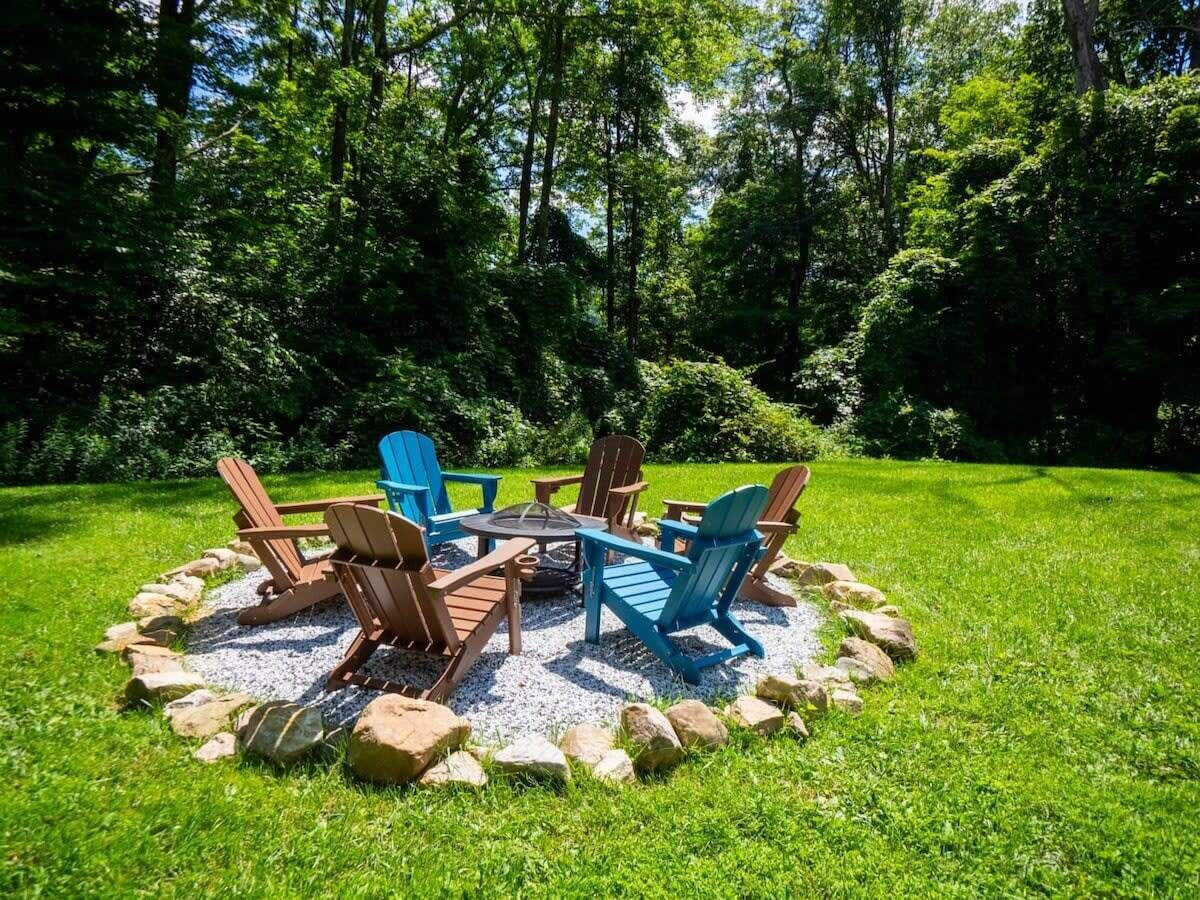 A circular outdoor seating area with Adirondack chairs and a fire pit, surrounded by a gravel ring and rocks, set in a lush green lawn.