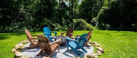 A circular outdoor seating area with Adirondack chairs and a fire pit, surrounded by a gravel ring and rocks, set in a lush green lawn.