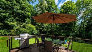 An outdoor deck with a grill, a glass table, and chairs, surrounded by a wooden deck and a grassy lawn. - Catamount, Zip Line, Private Setting, Book Now! (Egremont)