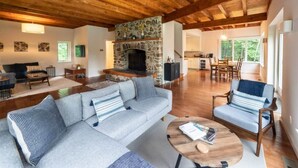A spacious living room with a stone fireplace, wooden ceiling beams, a sofa, a chair, and a dining area in the background. - Catamount, Zip Line, Private Setting, Book Now! (Egremont)