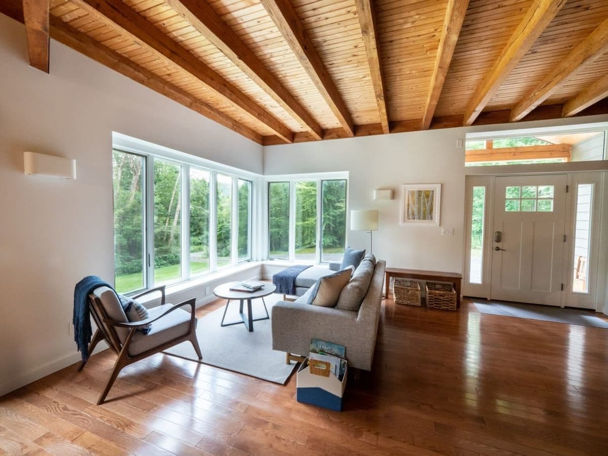 A spacious living room with hardwood floors, a sofa, a chair, and a coffee table. There are large windows providing natural light and a view of greenery outside.