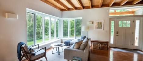 A spacious living room with hardwood floors, a sofa, a chair, and a coffee table. There are large windows providing natural light and a view of greenery outside.