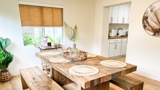 A wooden dining table with woven placemats and benches, a kitchen in the background, and a large potted plant on the left. - Boho Beach Villa- minutes to the ocean  (Vero Beach)