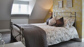 A bedroom with a sloped ceiling, a bed with floral bedding, a radiator, and a small table with a glass. - Belford House @ Seahouses (Seahouses)