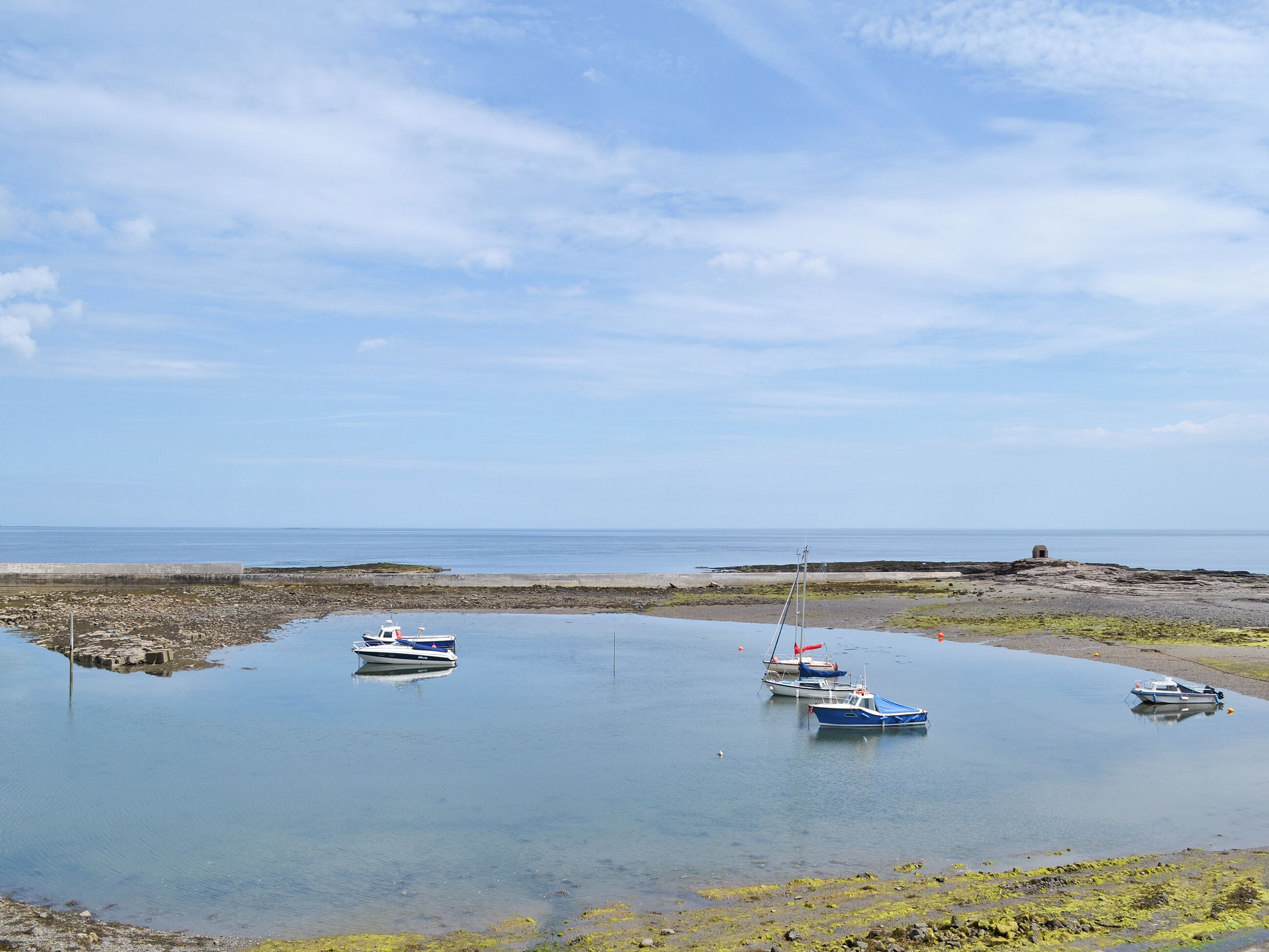 A calm harbor with several boats anchored near the shore, surrounded by rocky terrain and a clear sky.