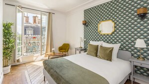 Une chambre d’hôtel avec un grand lit, un balcon donnant sur les immeubles, une chaise et une table.