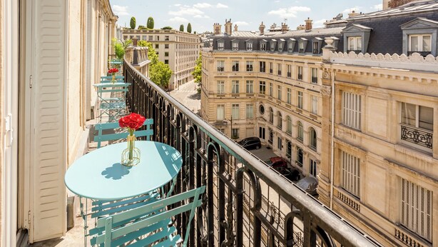 Un balcon avec une table ronde et des chaises, donnant sur un paysage urbain avec des bâtiments historiques.