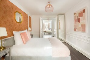 A hotel room with a large bed, a nightstand, a lamp, a mirror, and a hallway leading to another room. - Merveil - Family suite - Louvre - Victoires (Paris)