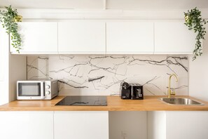 A kitchen with white cabinets, a marble backsplash, a microwave, a toaster, a sink with a gold faucet, and green plants on the counter. - Merveil - Family suite - Louvre - Victoires (Paris)