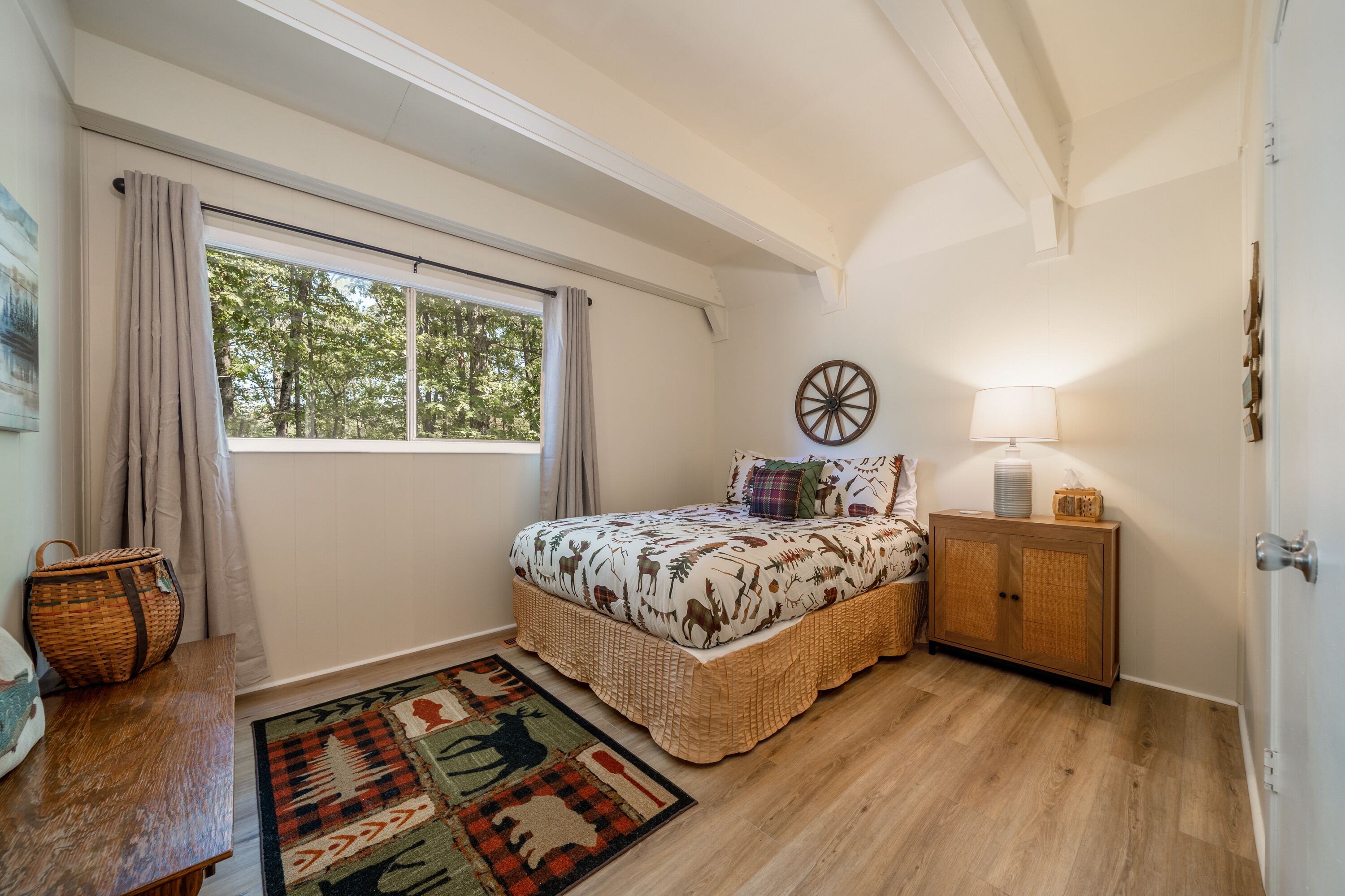 Une chambre avec une grande fenêtre, un lit recouvert d’une couvre-lit aux motifs naturels, une table de chevet avec une lampe et une armoire en bois.
