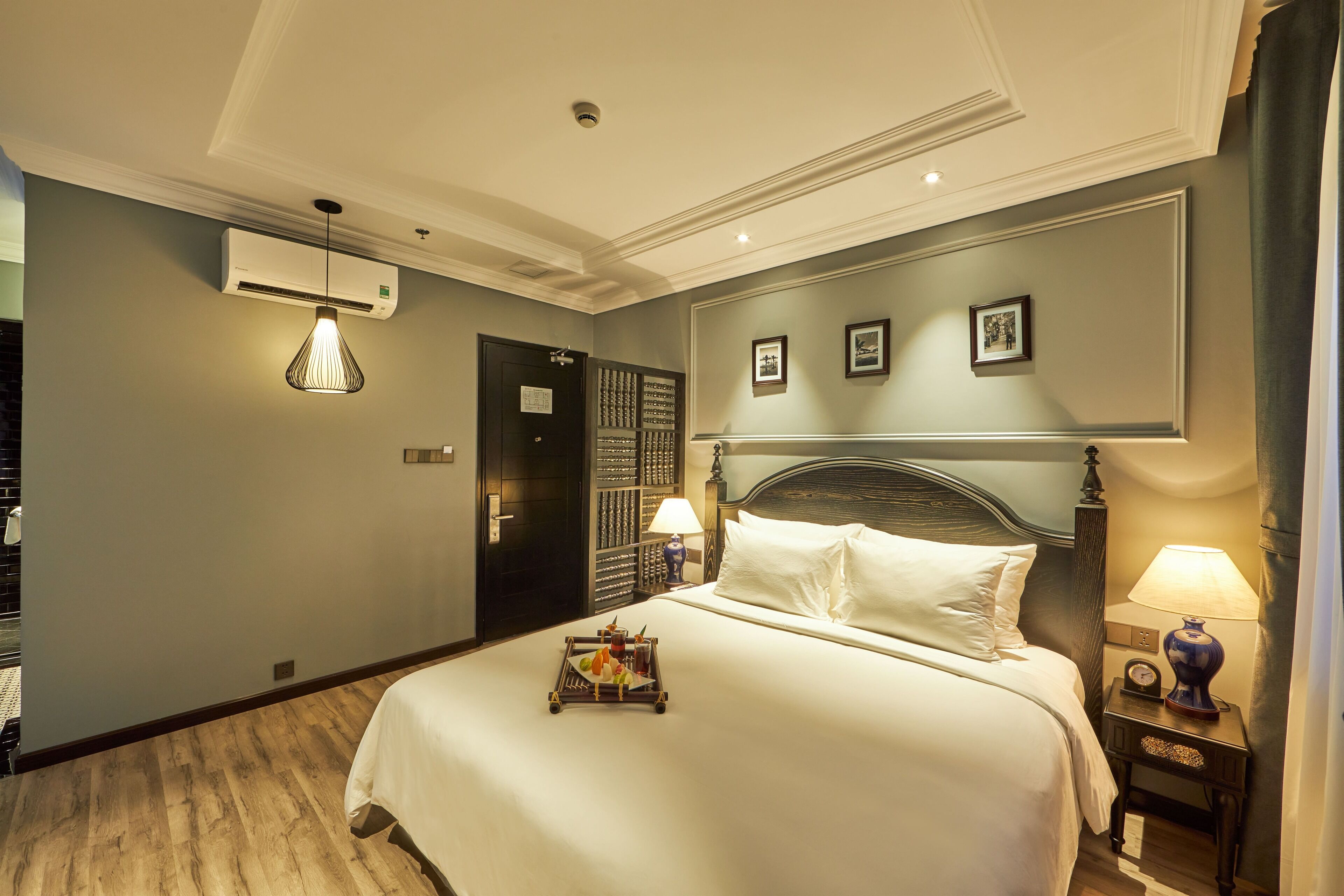 A hotel room with a neatly made bed, an air conditioning unit, a bedside table with a lamp, and a wall-mounted picture frame.