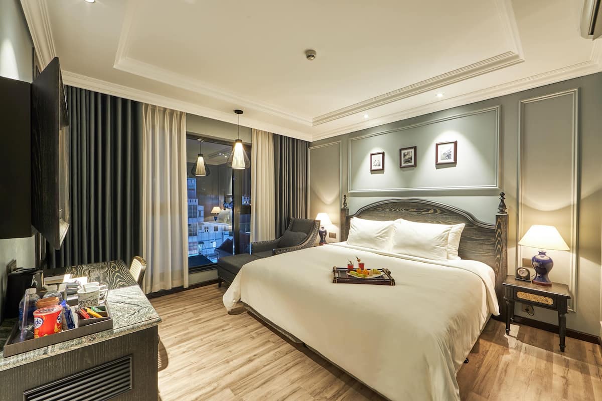 A hotel room with a large bed, a desk with a flat-screen TV, a chair, a nightstand with a lamp, and a window with curtains.