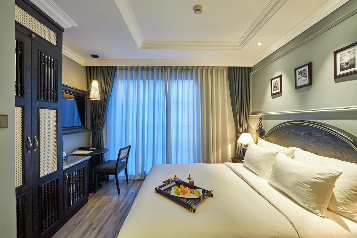 A hotel room with a bed, a desk, and a chair. There is a tray with fruits and a drink on the bed.