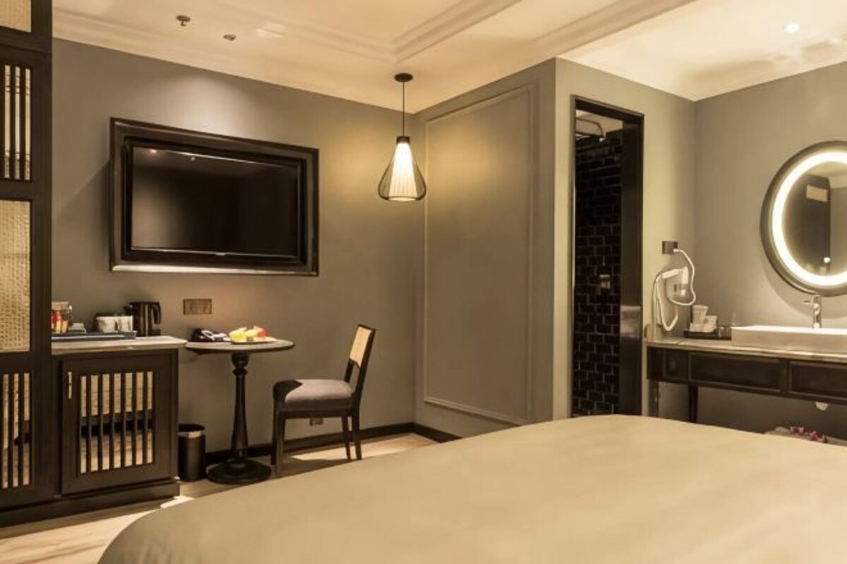 Ein Hotelzimmer mit Bett, Wandfernseher, kleinem Tisch mit Stuhl sowie einem Badezimmer mit Spiegel und Waschbecken.