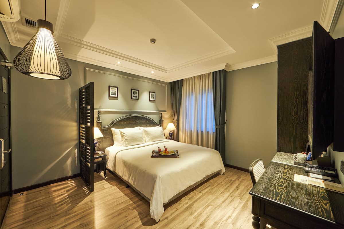 A hotel room with a bed, bedside table, lamp, and a desk with a computer and keyboard.