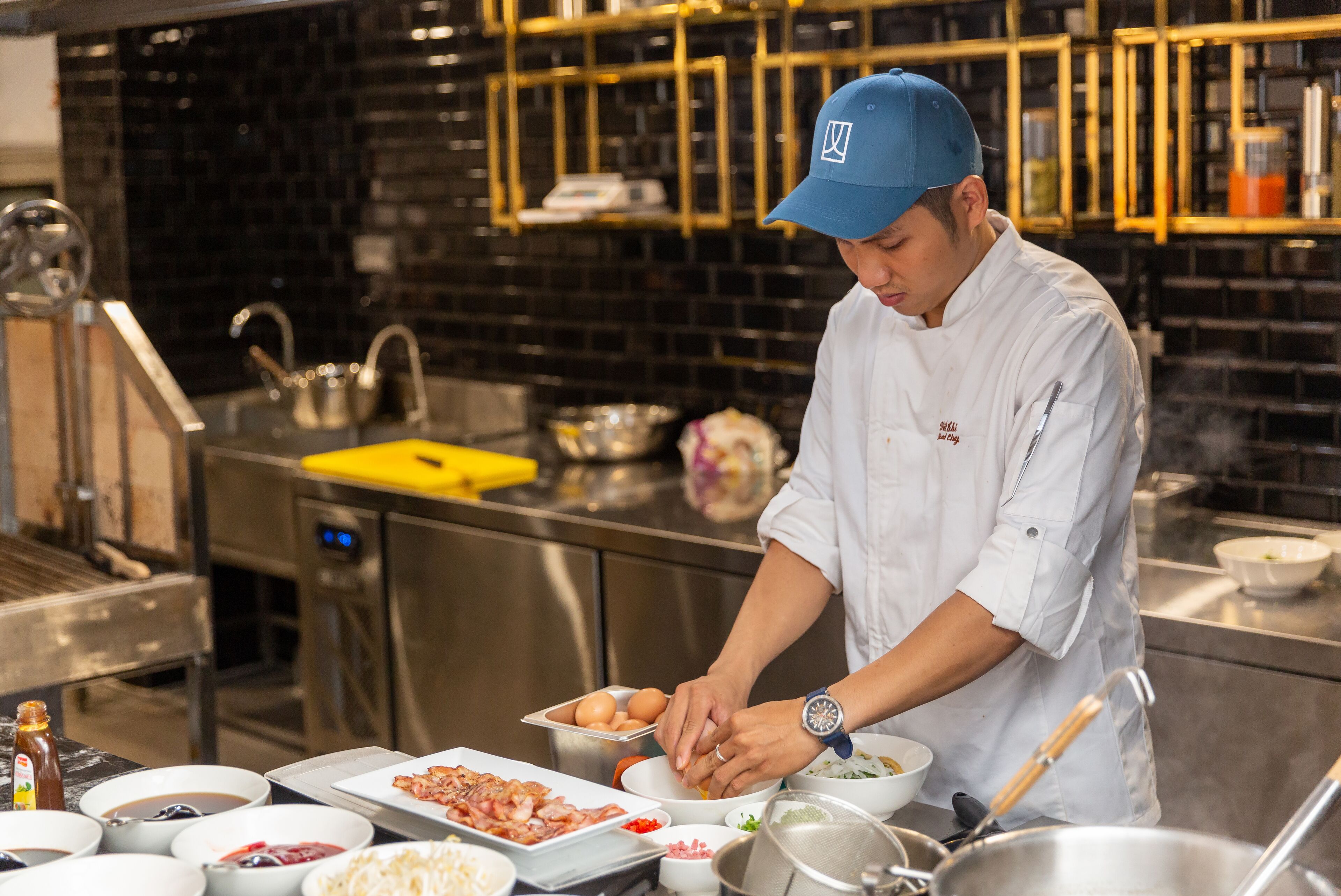 A chef preparing food in a modern kitchen with stainless steel appliances and black tiled backsplash.