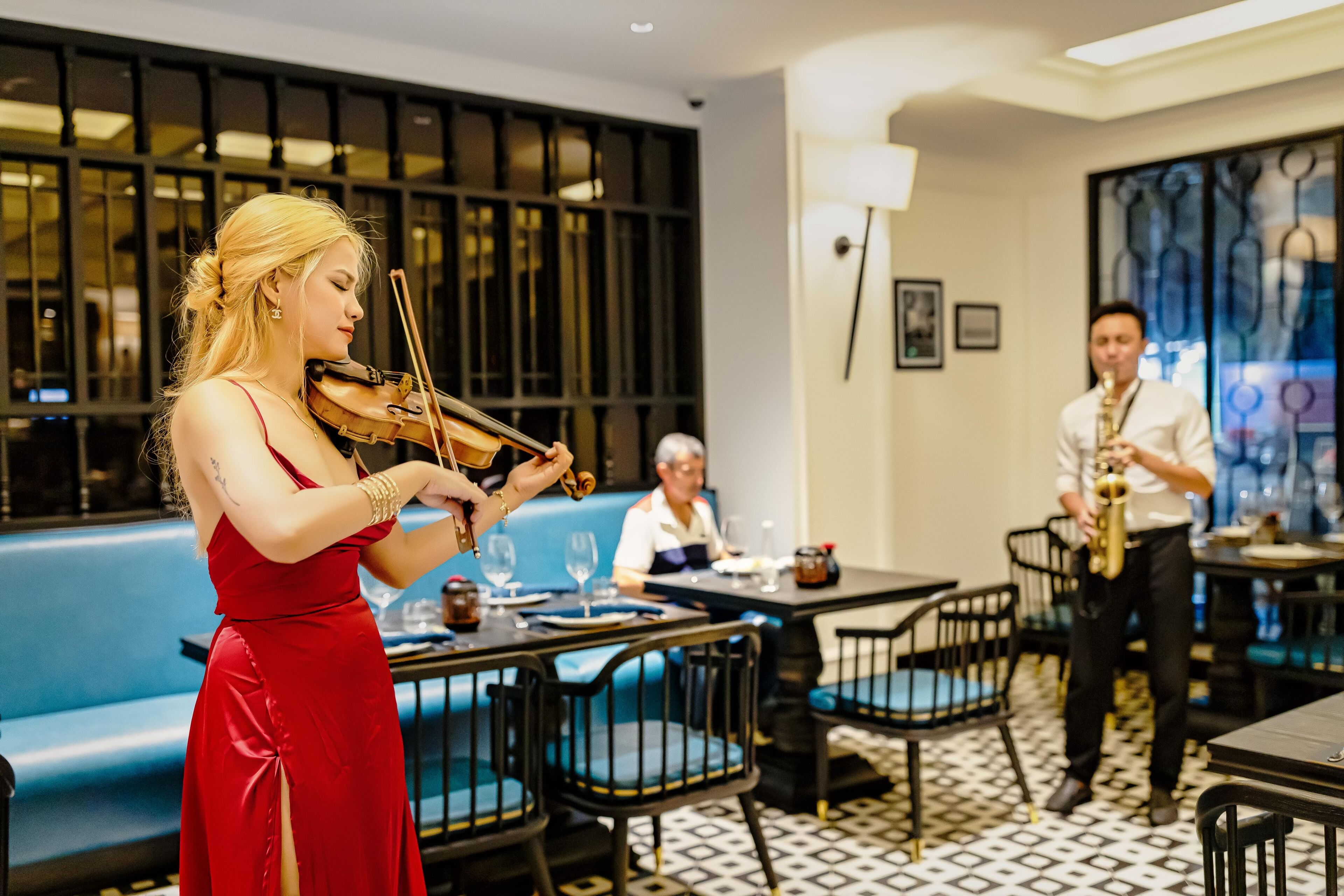 A woman playing the violin in a restaurant with a saxophonist and a seated patron.