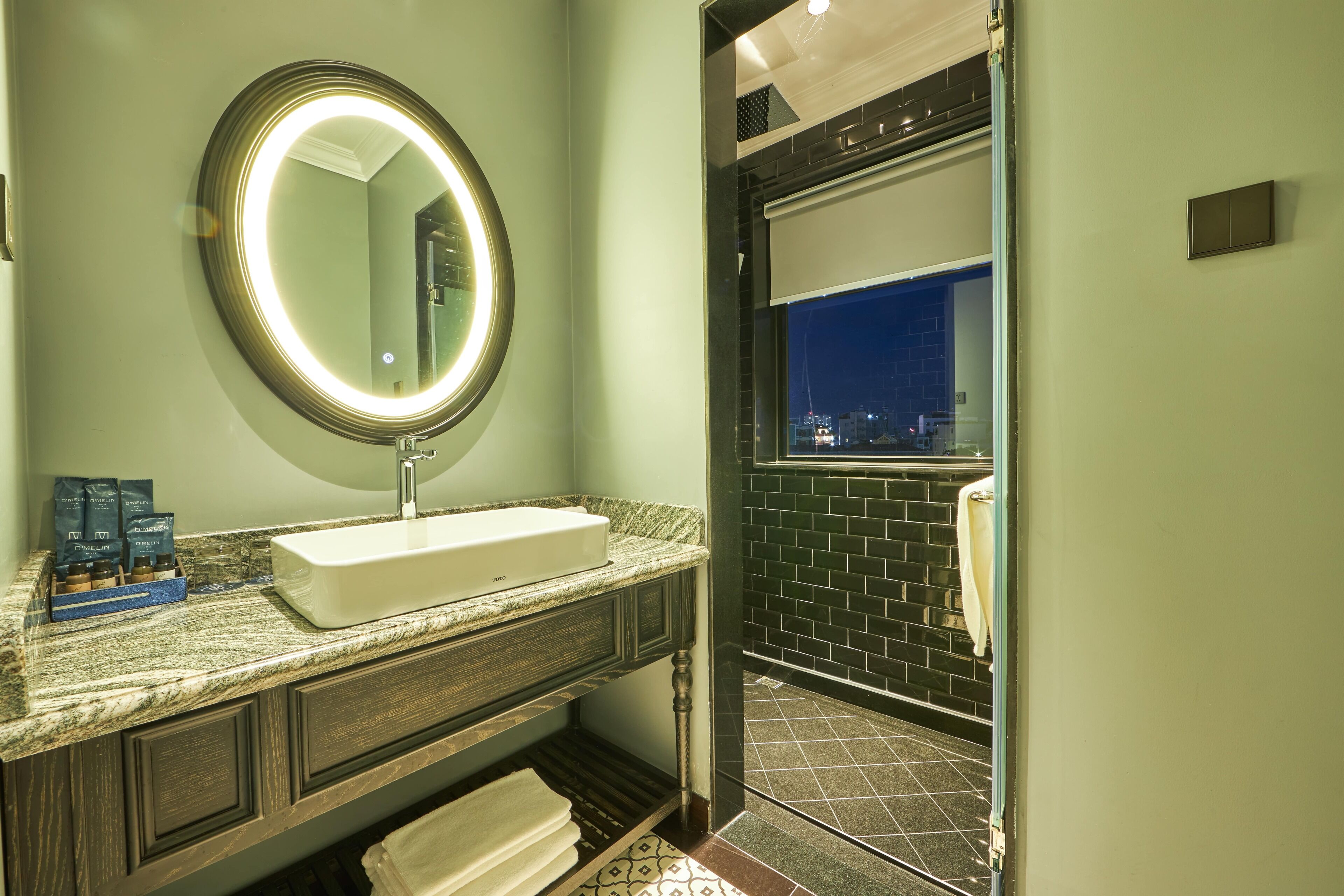 Une salle de bain avec un lavabo blanc, un miroir rond avec un éclairage intégré, un porte-serviettes et une vue sur la ville la nuit.
