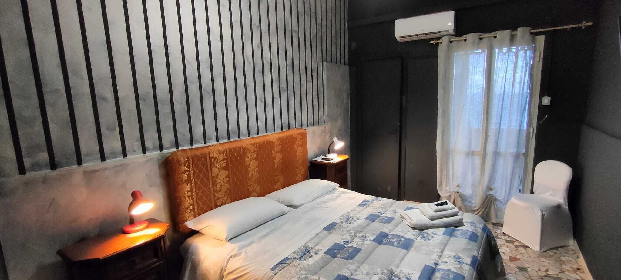Un dormitorio con una cama, dos mesitas de noche con lámparas, una silla, una ventana con cortinas y un equipo de aire acondicionado.