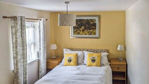 Un dormitorio con una cama, dos mesitas de noche, una lámpara colgante, una ventana con cortinas y un cuadro enmarcado en la pared.