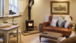 A cozy living room with a wood-burning stove, a leather sofa, a dining table, and a framed picture on the wall. - The Old Cobblers Cottage (Bakewell)
