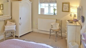 A bedroom with a large wardrobe, a bed with a pink floral bedspread, a wicker chair, a small table with a bowl and flowers, and a window with a decorative valance. - The Old Cobblers Cottage (Bakewell)