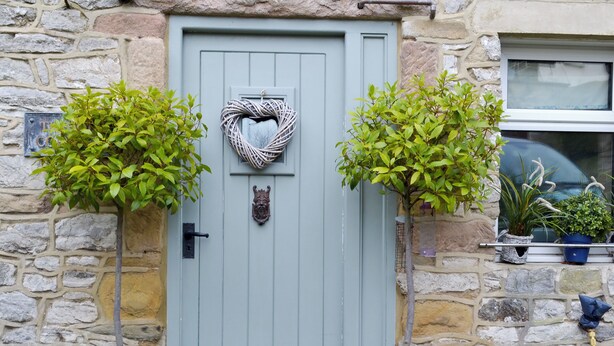 A stone building with a light blue door, two potted trees, and a small window with a plant. - The Old Cobblers Cottage (Bakewell)