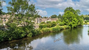A riverside view with stone buildings, lush greenery, and ducks swimming. - The Old Cobblers Cottage (Bakewell)