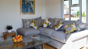 A living room with a grey sofa, a glass coffee table, a painting on the wall, and a view through large windows. - Y Bwthyn at Tyddyn Obry (Ty Croes)