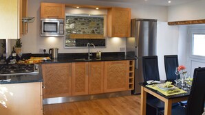 A modern kitchen with wooden cabinets, a built-in microwave, a stainless steel refrigerator, and a dining area with a table set for two. - Y Bwthyn at Tyddyn Obry (Ty Croes)