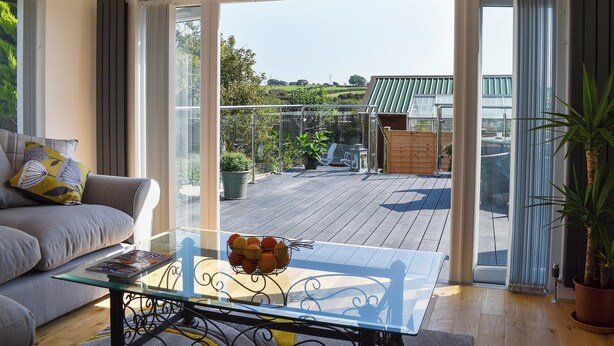 A modern living room with a glass table, a sofa, and a view of a wooden deck and greenery outside. - Y Bwthyn at Tyddyn Obry (Ty Croes)