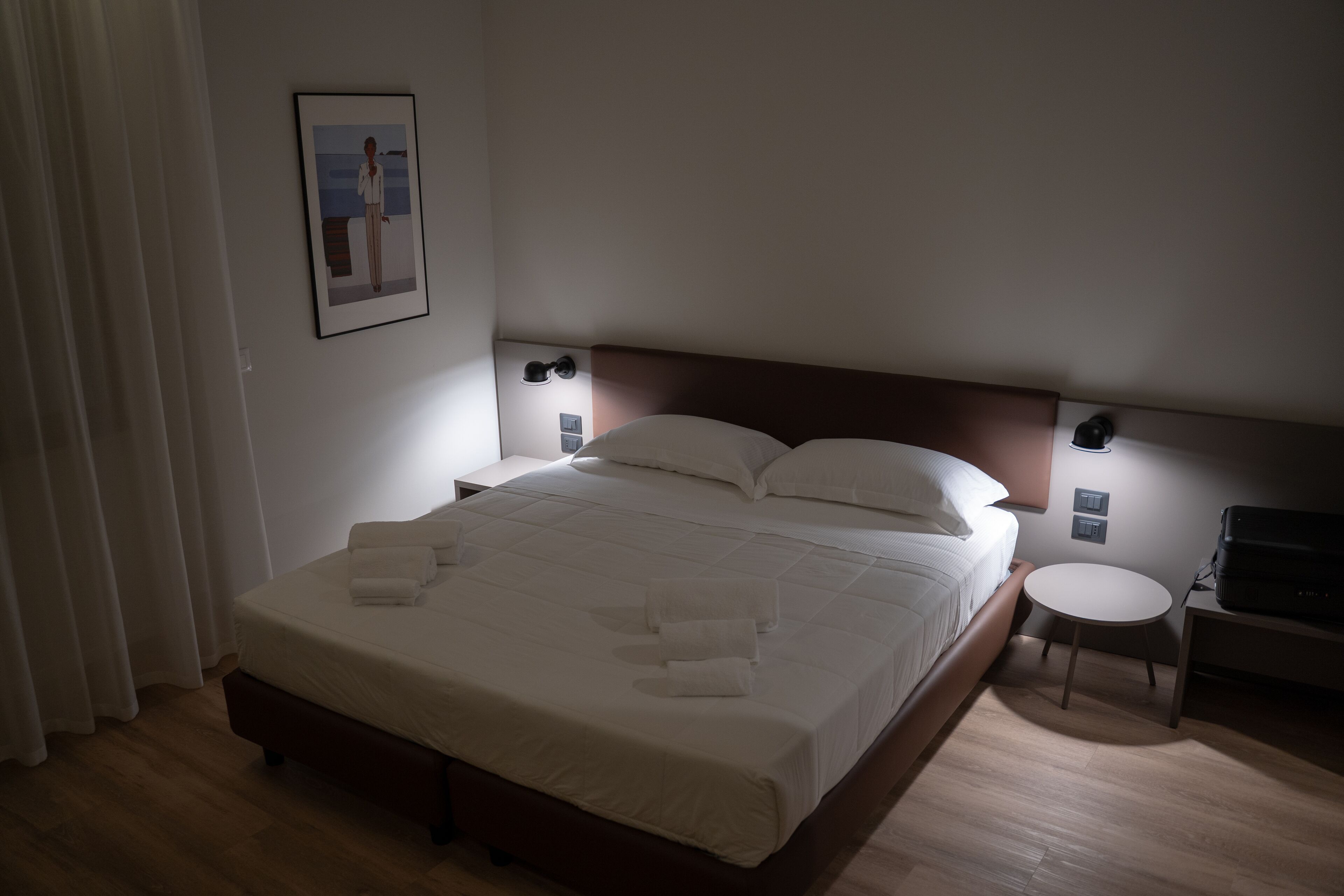 Una cama bien tendida con sábanas y toallas blancas, una mesita de noche con lámpara, y una pequeña mesa redonda con una computadora portátil.