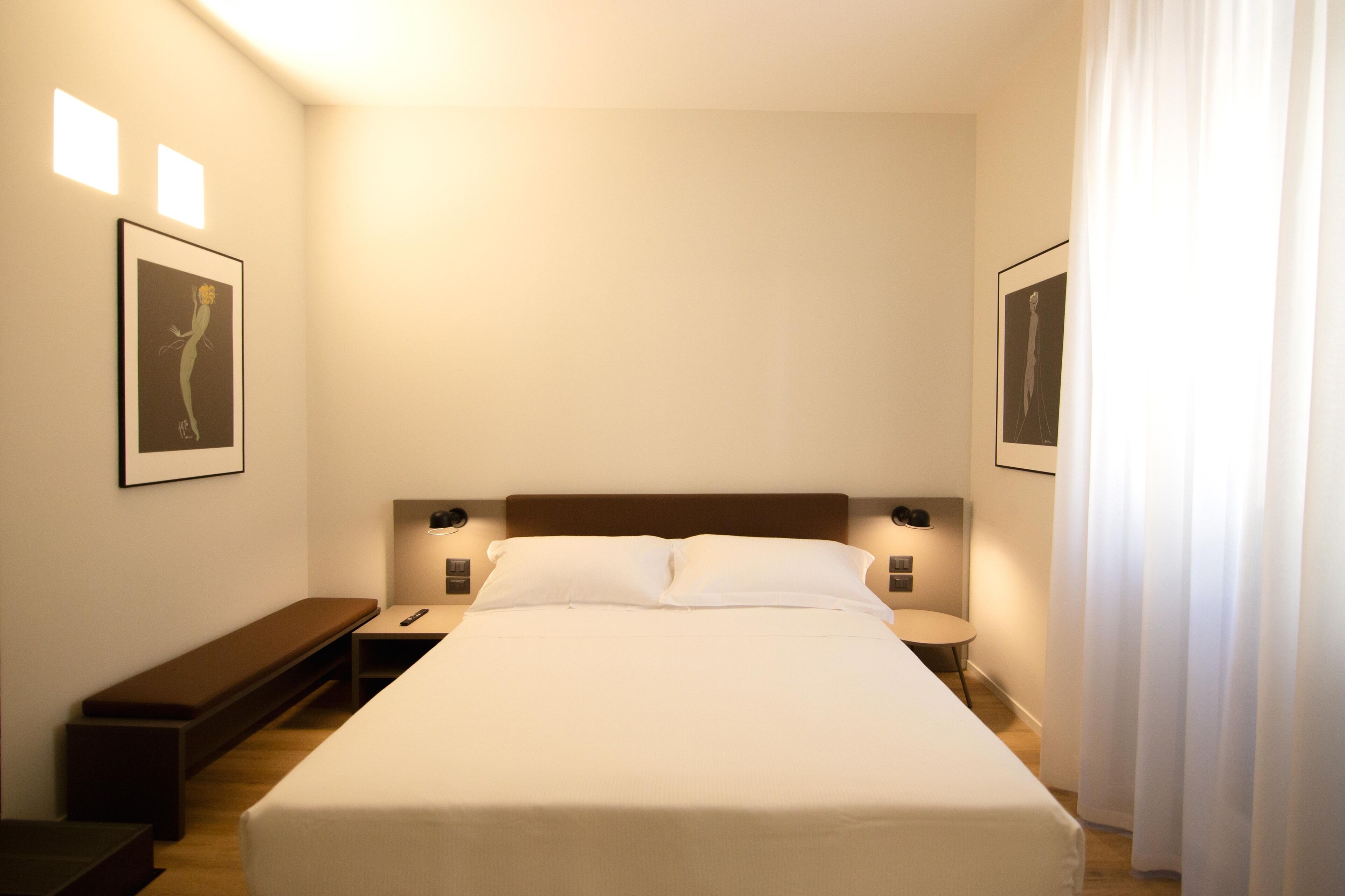 Una habitación de hotel ordenada con una cama grande, mesitas de noche, un banco y cuadros en las paredes.
