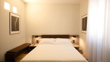 Una habitación de hotel ordenada con una cama grande, mesitas de noche, un banco y cuadros en las paredes.