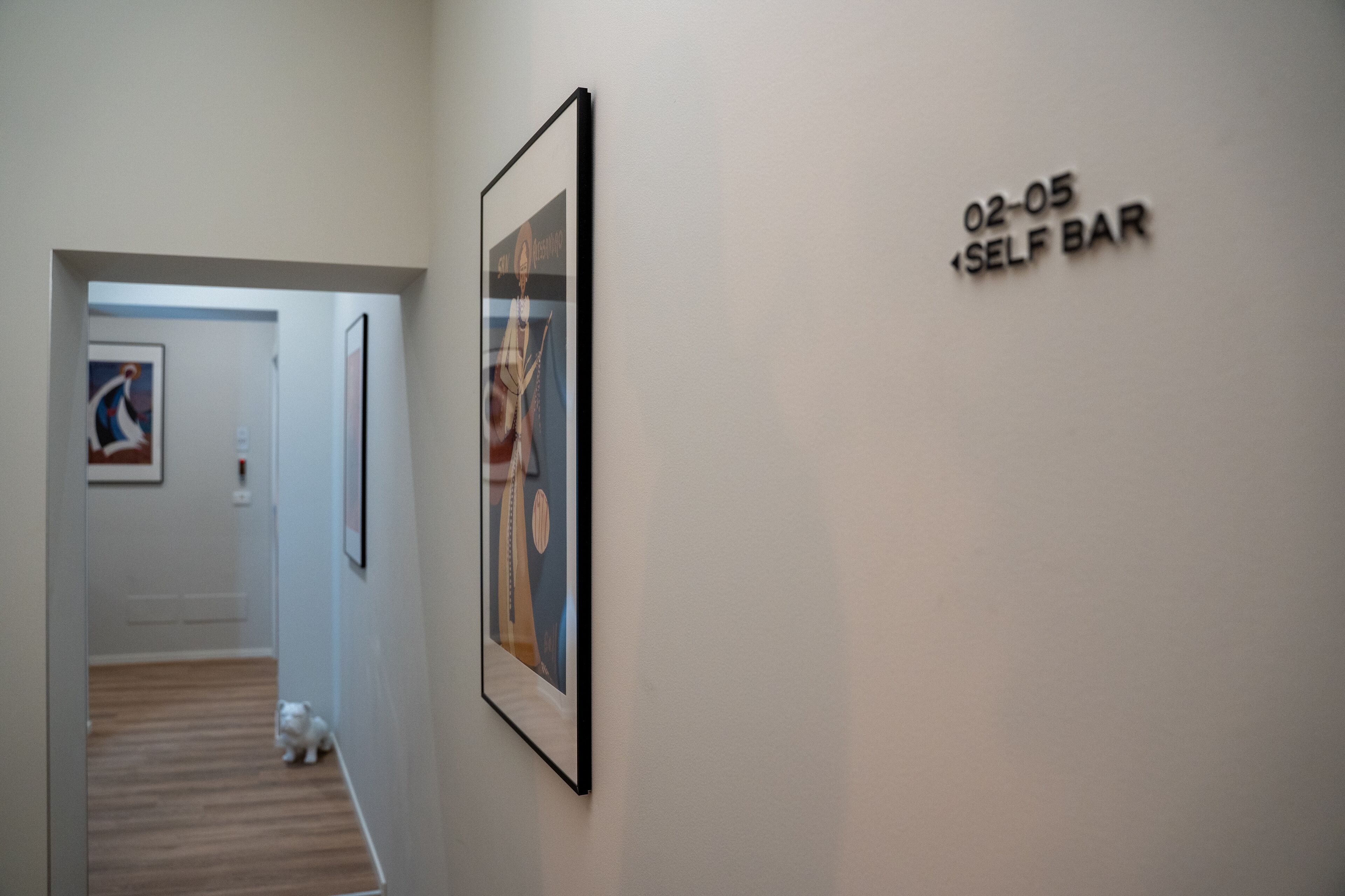 A hallway with a framed picture on the wall, a small dog on the floor, and a sign reading "02-05 SELF BAR".