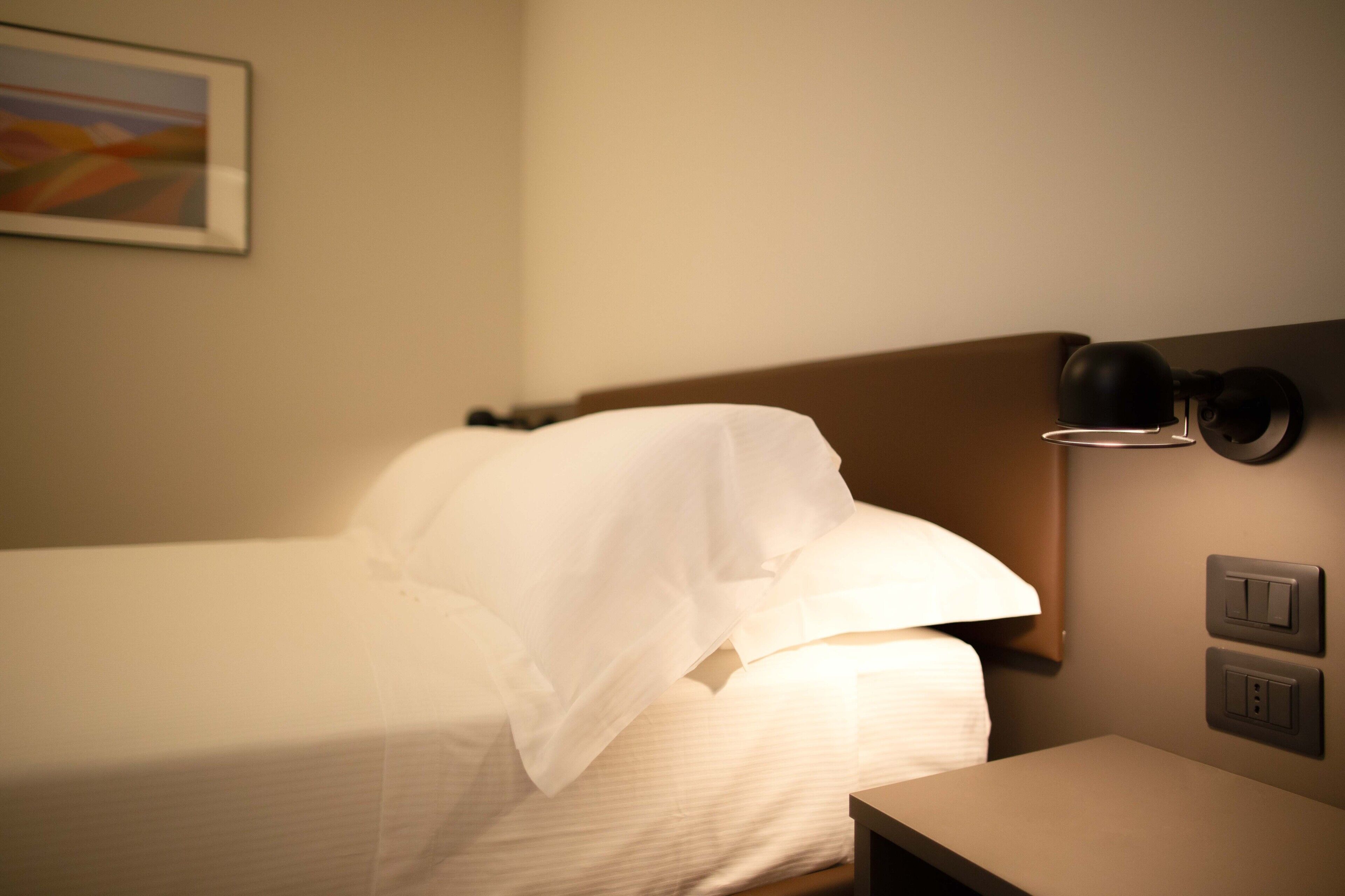 A hotel room with a neatly made bed, a bedside table, a wall-mounted lamp, and a painting on the wall.