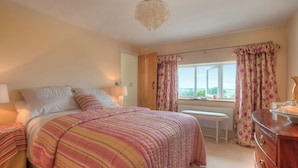 Une chambre à coucher comprenant un lit, des tables de chevet, une commode, une fenêtre avec des rideaux et une vue sur l’extérieur.
