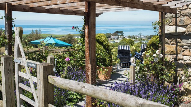 Une pergola en bois, adossée à un mur de pierre, offrant une vue sur la côte, avec des maisons et un parasol bleu.