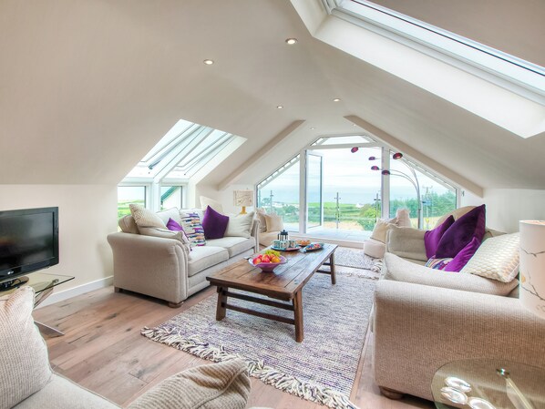 A spacious loft with a high ceiling, skylights, a flat-screen TV, and a large window offering a view of the outdoors.