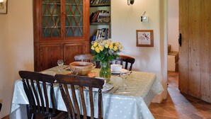 A dining area with a wooden table, chairs, a vase with flowers, and a bookshelf in the background.
